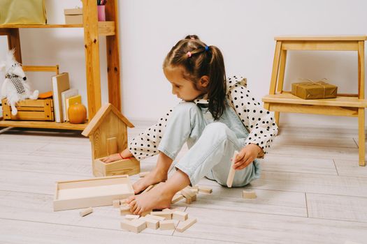 A girl with two ponytails on her head sits on the floor in her room and plays with wooden cubes. The girl sits on the floor and builds a tower out of the constructor.