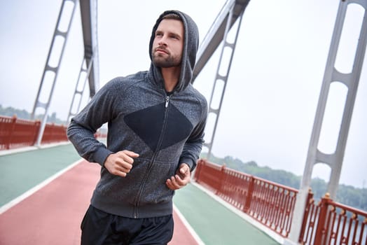 Morning workout. Close up of young man in sports clothing exercising while jogging on the bridge outdoors