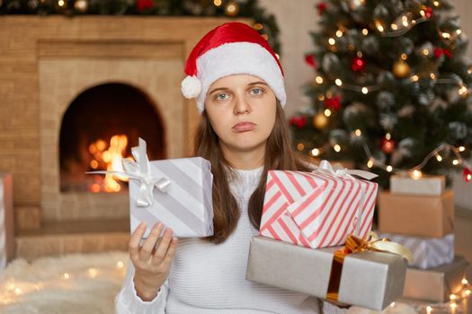 Young pretty girl is not satisfied with her gifts, posing near fireplace and x-mas tree with present boxes in hands, looking at camera with upset expression, wearing santa claus hat and white sweater