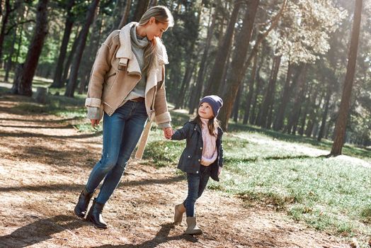 A little curly girl in hat is walking with her mother in woods. Cold season, bright sun is seen through the trees