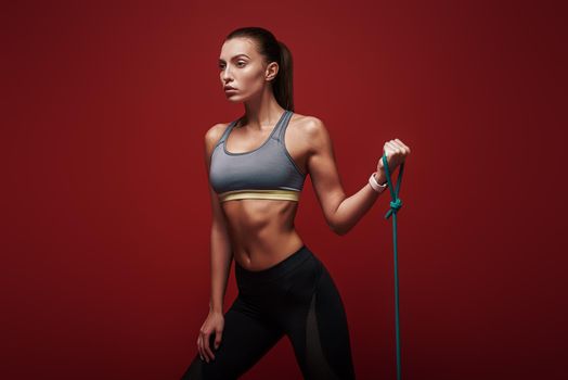 Self-determined woman does exercises with rubber band, works on hands and legs, wears comfortable sportsclothes, poses over red background. Sport and goals concept