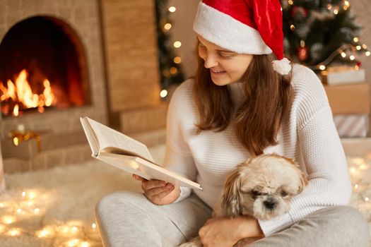 Smiling young woman relaxing near fireplace, reading book in cozy christmas atmosphere with decorated xmas tree in background, lady looking at pages with concentrated look, hugging her dog.