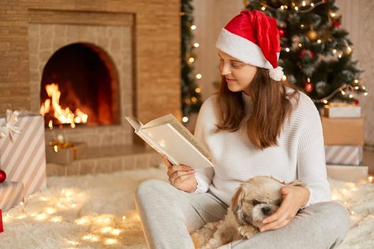 Positive young woman enjoying to spend time with her dog, adorable female posing in festive living room with her pet, reading book while sits near Christmas tree and fireplace.