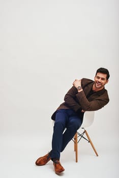 Full-length portrait of stylish dark-haired man wearing blue trousers and brown jacket, sitting on a white chair, leaning back and laughing isolated over white background