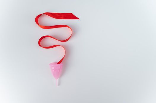 Pink menstrual cup and red satin ribbon on a white background. Copy space.