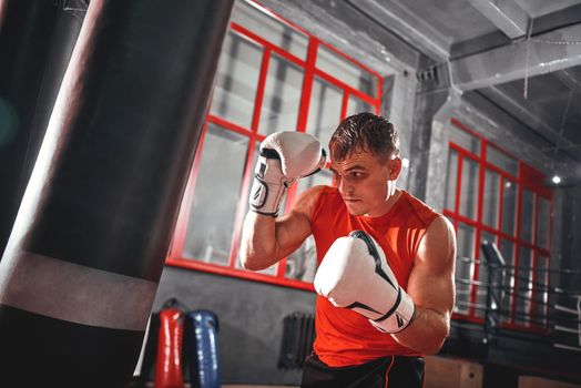 Confident muscular sportman in sports clothing hard training on heavy punch bag. Young boxer with white boxing gloves in safeguard stand on red window background