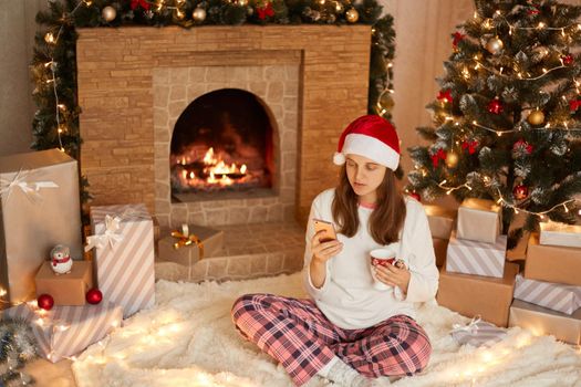 Young woman wearing santa hat and pajamas sitting on floor amongst wrapped christmas presents, fireplace and xmas tree, drinking hot tea and shopping online on her phone.