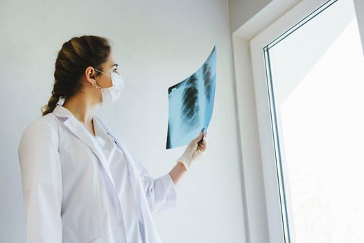 A radiologist in a medical mask and gloves radiates an x-ray image of the patient's lungs. The girl doctor works on the front line. The doctor is holding an X-ray of his lungs.