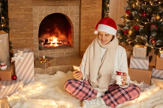 Photo of positive lady sitting with cellphone and holding beverage mug in house indoors with christmas x-mas ornament, wearing santa hat, scarf and checkered pants, having video call.