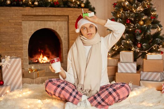 Photo of cute young ill girl holds thermometer in mouth and sore throat spray in hands, drinking hot beverage, wears santa cap, white pullover and checkered pants, posing in decorated living room.