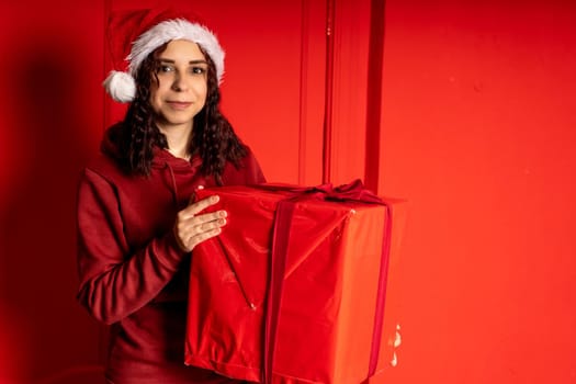 Young woman in Santa Claus hat holds big gift, standing near red wall. Happy female in Christmas hat with box of gift. Concept of holidays, presents and good mood