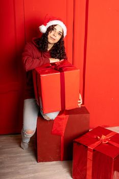 Young attractive woman in Santa Claus hat sits on gifts near red wall. Smiley female sitting on big box of gift. Concept of holidays, gifts and good mood