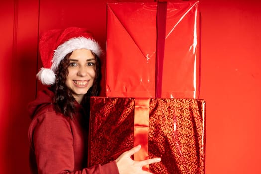 Young woman in Santa Claus hat holds big gifts, standing near red wall. Happy female in Christmas hat with boxes of gift. Concept of holidays, presents and good mood