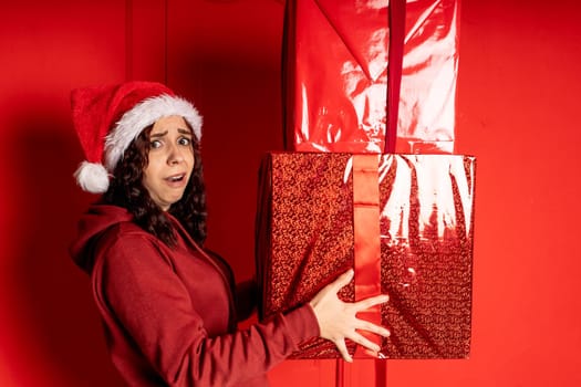 Young woman in Santa Claus hat holds heavy big gifts, standing near red wall. Exhausted female in Christmas hat with boxes of gift. Concept of holidays and presents