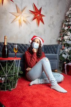 Young woman in medical mask and Santa Claus hat sitting on floor near sofa in room. Alone charming brunette in protective mask celebrating of safe Christmas during coronavirus pandemic