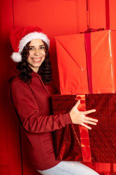 Young woman in Santa Claus hat holds big gifts, standing near red wall. Happy female in Christmas hat with boxes of gift. Concept of holidays, presents and good mood