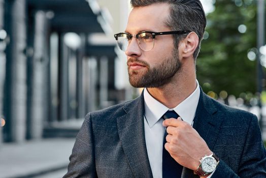 Close-up handsome and successful man in spectacles and in an expensive suit. He is in a white shirt with a tie. The man straightens his tie, his face unshaven.