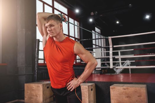 Focused young muscular athlete in sports clothing stretching with expander while warming up before training in black boxing gym