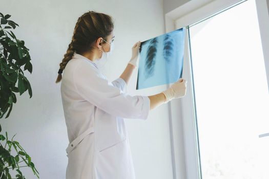 A radiologist in a medical mask and gloves radiates an x-ray image of the patient's lungs. The girl doctor works on the front line. The doctor is holding an X-ray of his lungs.