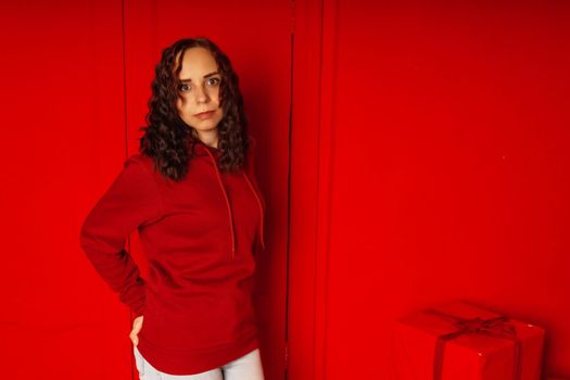 Young woman in hoodie on red background. Curly brunette poses near red wall