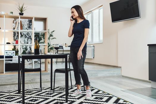 Dark-haired, sad and wasted alcoholic woman standing in the kitchen, talking on the phone at home. She is completely drunk, looking depressed, lonely and suffering hangover in alcoholism and alcohol abuse. Full-length shot