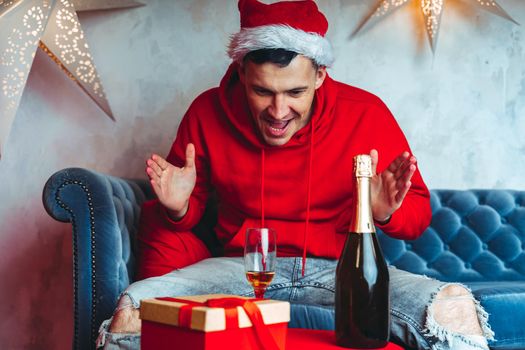 Young man in Santa hat celebrating Christmas in solitude at home. Adult guy rests, sitting on sofa in room. Concept of holidays and good mood