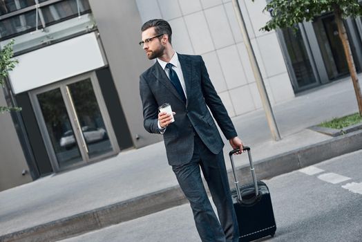 A young businessman spectacled drinks coffee and crosses the road. In his right hand he holds a suitcase on wheels. He looks at the road