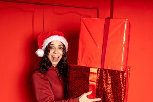 Young woman in Santa Claus hat holds big gifts, standing near red wall. Happy female in Christmas hat with boxes of gift. Concept of holidays, presents and good mood