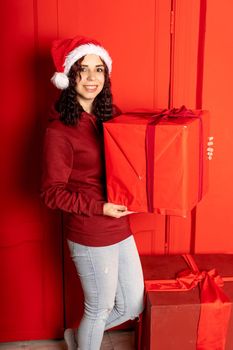 Young woman in Santa Claus hat holds big gift, standing near red wall. Happy female in Christmas hat with box of gift. Concept of holidays, presents and good mood