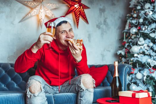 Young man in santa hat drinks champagne and eats pizza, sitting on sofa in room. Happy male resting with food and alcohol. Concept of Christmas celebration at home