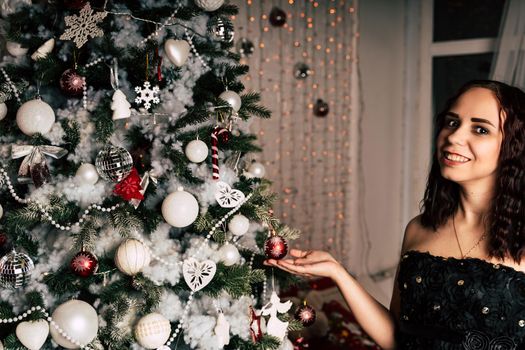 Portrait of charming brunette in black dress near Christmas tree. Young attractive woman posing at coniferous tree with decorative adornments. Concept of Christmas celebration at home