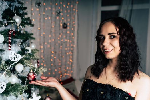 Portrait of charming brunette in black dress near Christmas tree. Young attractive woman posing at coniferous tree with decorative adornments. Concept of Christmas celebration at home