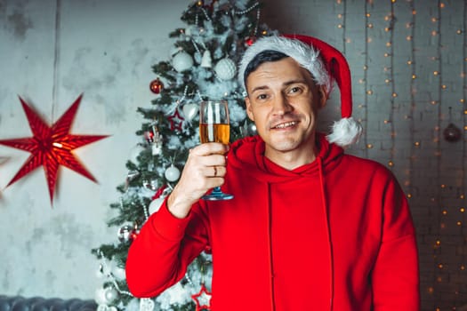 Young man in santa hat with glass of champagne on background of Christmas tree. Adult male posing at coniferous tree with decorative adornments. Concept of Christmas celebration at home