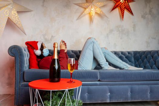 Close up of bottle and glass of champagne on table. Woman in medical mask and Santa Claus hat browses smartphone, lying on couch in background Concept of safe Christmas celebration during coronavirus