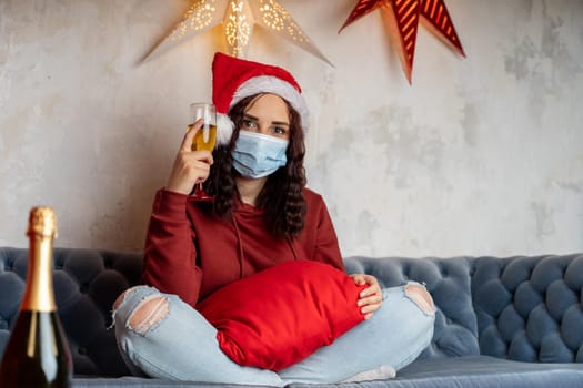 Young woman in medical mask and Santa Claus hat offers toast, looking at camera. Alone female sitting on couch and celebrating of safe Christmas during coronavirus pandemic