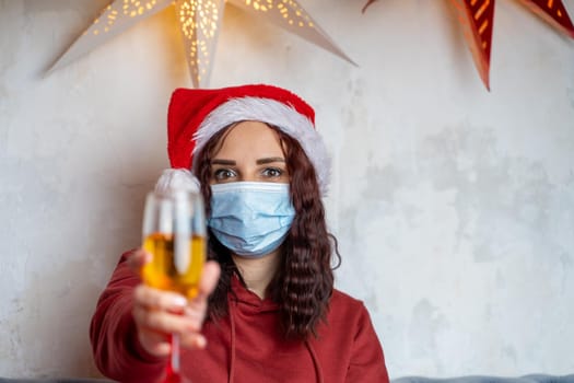 Close up of young woman in medical mask holds glass of champagne. Brunette in protective mask offers toast, celebrating of safe Christmas during coronavirus pandemic