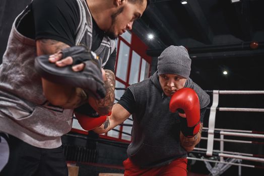 Training hard. Muscular athletic tattooed man in sports clothing training on boxing paws with partner opposite boxing ring
