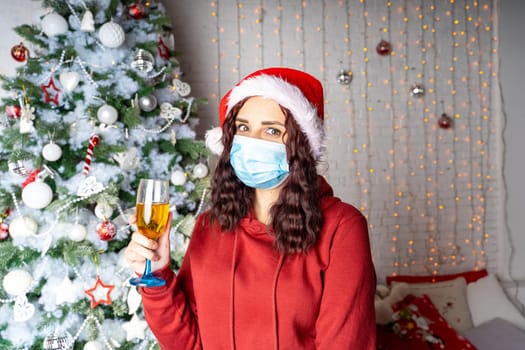 Young woman in medical mask and santa hat with glass of champagne on background of Christmas tree. Charming brunette in protective mask celebrating of safe Christmas during coronavirus pandemic