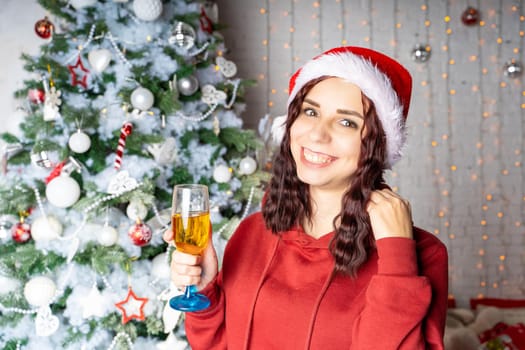 Young woman in Santa Claus hat with glass of champagne standing near Christmas tree. Charming brunette posing at coniferous tree with decorative adornments. Concept of Christmas celebration at home