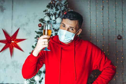 Young man in medical mask with glass of champagne on background of Christmas tree. Adult male celebrating of safe Christmas during coronavirus pandemic