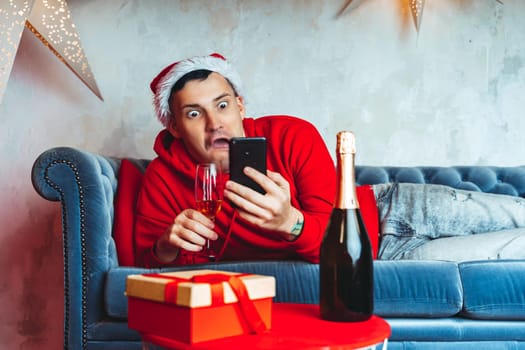 Young handsome man in Santa Claus hat with glass of champagne taking selfie on smartphone. Funny guy photographing himself on mobile phone and lying on couch. Concept of Christmas celebration at home