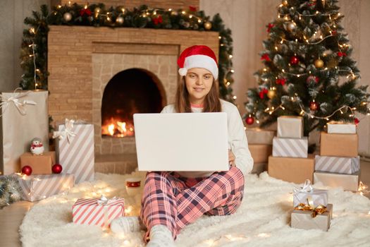 Young woman doing Christmas shopping online or having video call while sitting on floor near fir tree with her laptop, looking at screen with glad expression, wearing casual attire and santa hat.