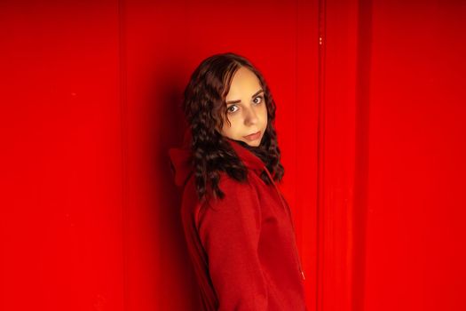Young woman in hoodie on red background. Curly brunette poses near red wall