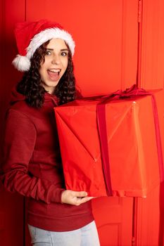 Young woman in Santa Claus hat holds big gift, standing near red wall. Happy female in Christmas hat with box of gift. Concept of holidays, presents and good mood