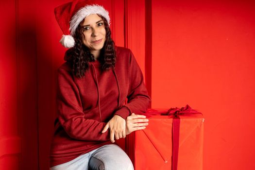 Young attractive woman in Santa Claus hat sits on gifts near red wall. Smiley female sitting on big box of gift. Concept of holidays, gifts and good mood