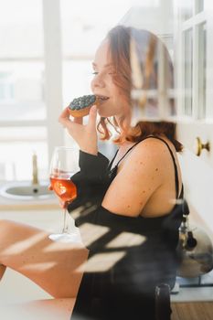 A young girl in a black negligee is sitting in the kitchen eating a cake and drinking wine. Bright kitchen. Sexy girl is resting. Double exposure and glare.