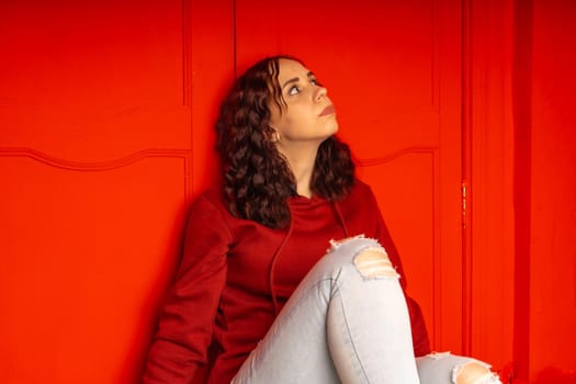 Young woman sitting on floor. Curly brunette poses near red wall