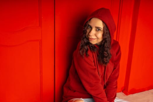 Young woman in hood sitting on floor. Curly brunette poses near red wall