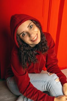 Young woman in hood sitting on floor. Curly brunette poses near red wall