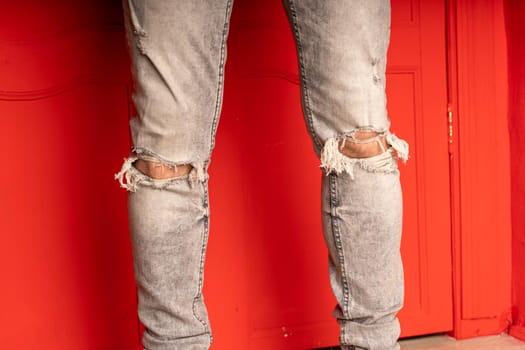 Close up of men's legs in jeans with holes on red background. Fashionable holes in grey jeans on unrecognizable man